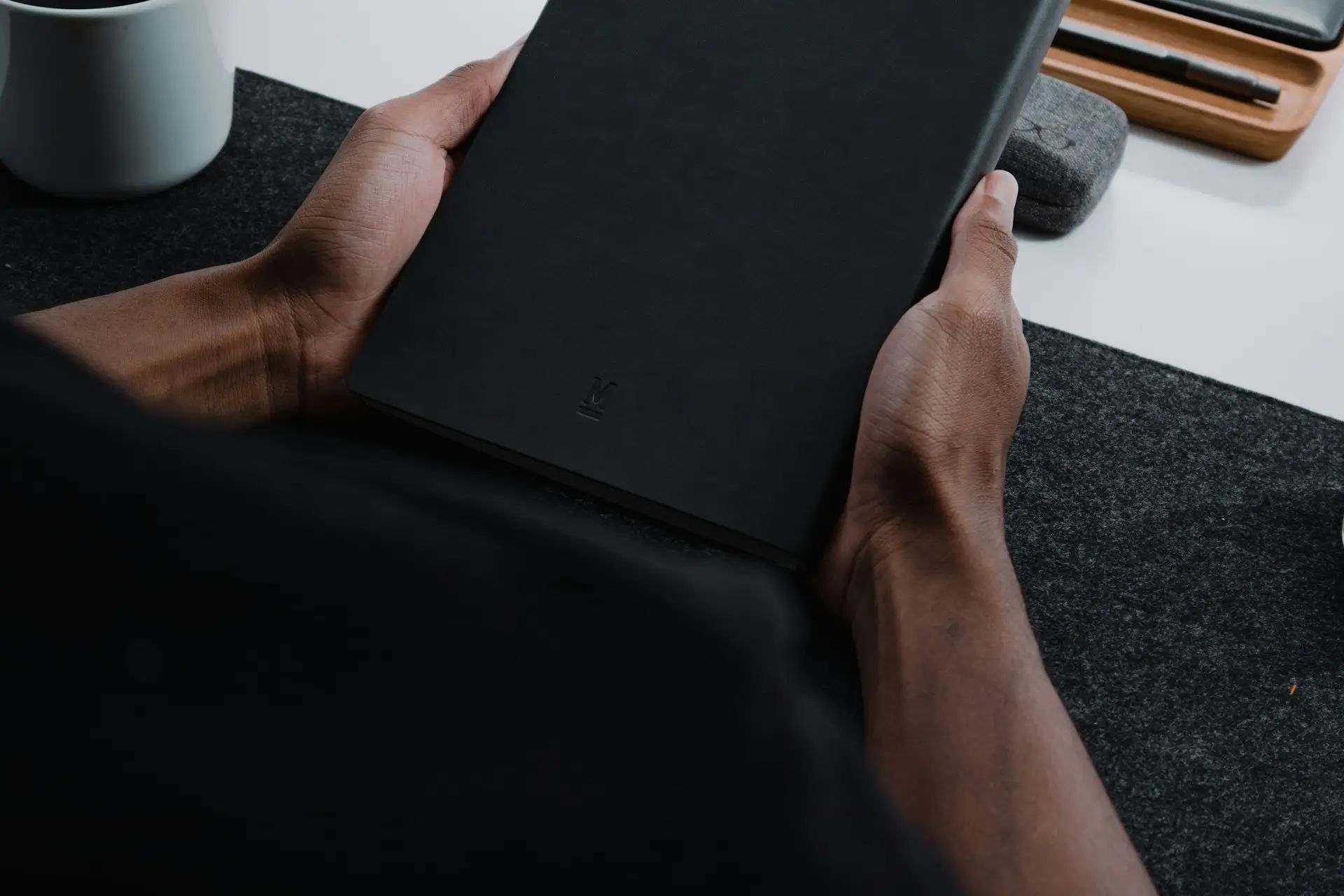 A close-up of a person's hands holding a sleek black notebook, set against a dark felt surface. A white coffee cup and a wooden organizer with pens are visible in the background, creating a minimalist workspace atmosphere.